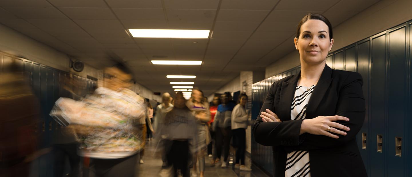 A woman standing with her arms folded across her chest, in a hallway of a school with blue lockers along the wall and people walking behind her.