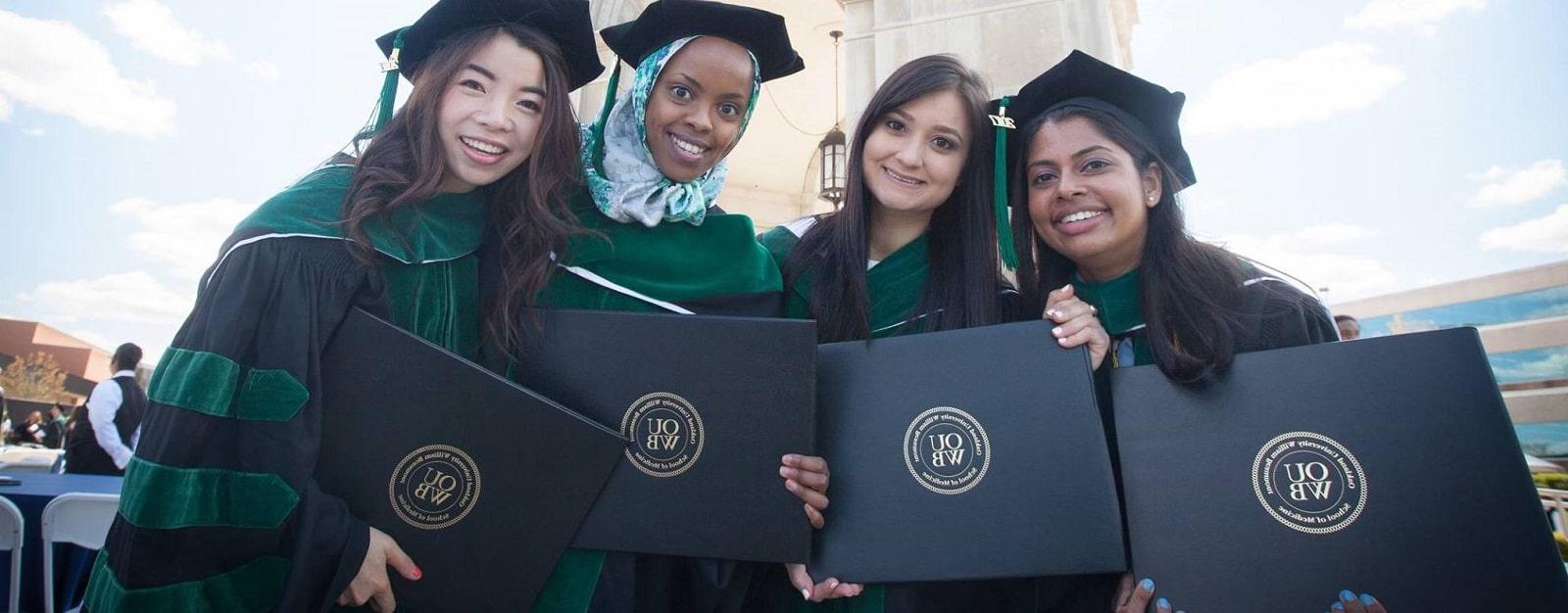 四名应届毕业生戴着帽子，身穿长袍，手持毕业证书站在艾略特塔前.