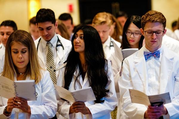 Students stand, holding programs, at the white coat ceremony.