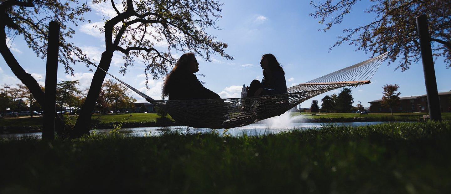 Two people seated, facing each other, in a hammock with a lake and fountain behind them.