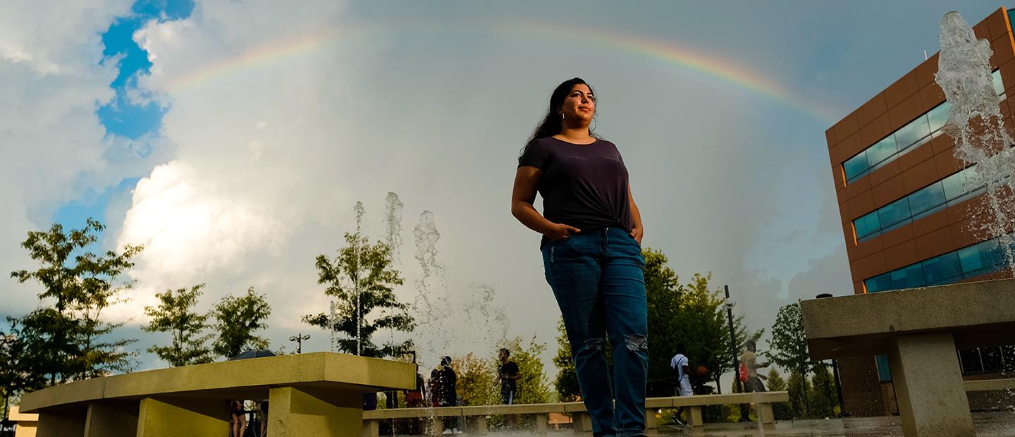 一位年轻女子站在365英国上市官网的校园里，双手插在口袋里, 身后有彩虹和喷泉.